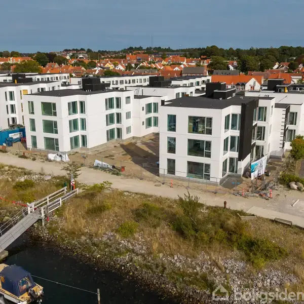 Flere moderne, hvide lejlighedsbyggerier står stille nær en havn med små både. I baggrunden ses en landsby med røde hustage omgivet af grønne områder.