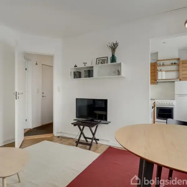 Et lille, lyst opholdsrum med hvidmalede vægge indeholder et TV på et bord, placeret mod væggen. Rummet har en dør mod gangen og en åben adgang til et køkken med komfur og køleskab. Møblerne inkluderer et rundt spisebord og små borde.