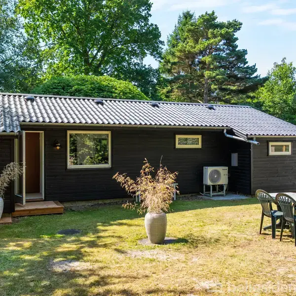 Et sort træhus står stille i en frodig have. Ved siden af huset står et bord med fire stole. Omkring er der grønne træer og buske.