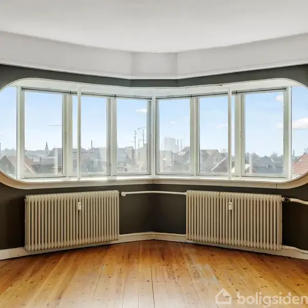Store vinduer med hvid ramme i et hjørne, indrammer en udsigt over byens tage. Under vinduerne er der to radiatorer. Rummet har brune vægge og et trægulv.