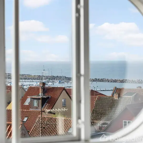 Et vindue indrammer udsigten over rødtage og en skov ved vandet i baggrunden under en blå himmel.