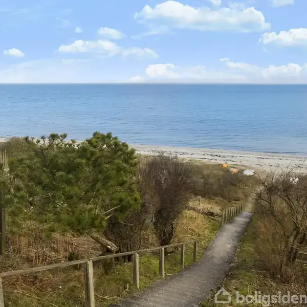 En sti fører ned mod en sandstrand med græs og buske omkring. Foran ligger havet under en blå himmel med lette skyer.