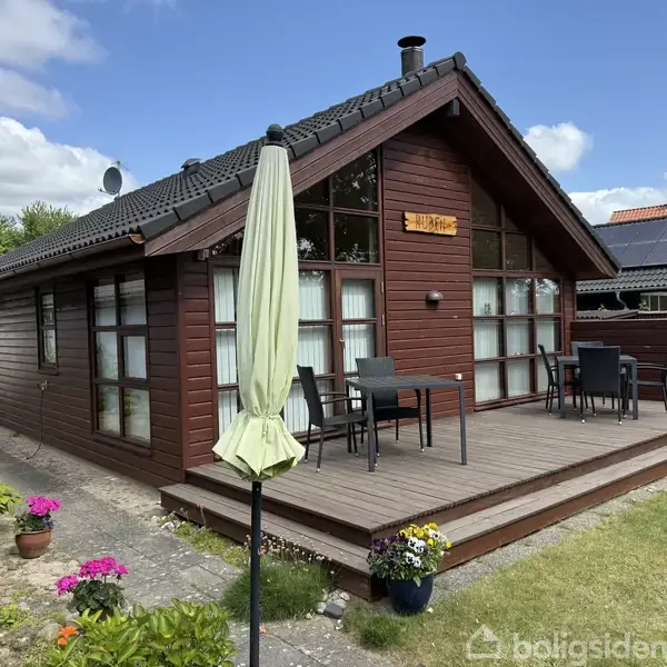 Træhus med højt tag, placeret i en have med blomster og havebord på træterrasse. En skilt med teksten ”Starlight” er fastgjort på gavlen. Solpaneler ses på nabotaget.