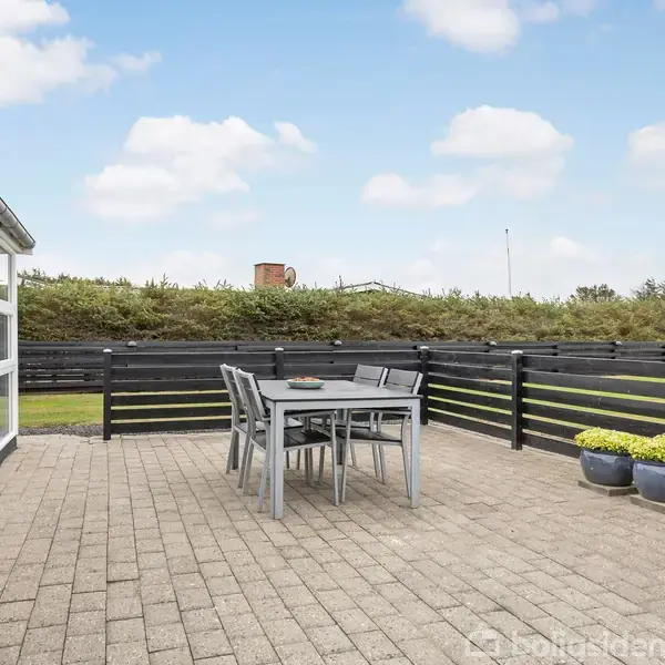 Terrasse med et bord og seks stole står på en flisebelagt overflade. Omkranset af et sort stakit og grøn beplantning, ses en åben himmel med spredte hvide skyer i baggrunden.