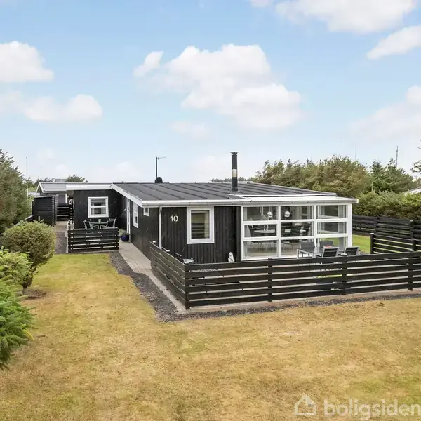 Et sort træhus med nummer 10 står placeret på en velplejet græsplæne omgivet af en sort stakit, mens buske og træer danner baggrund mod en blå, skyet himmel.