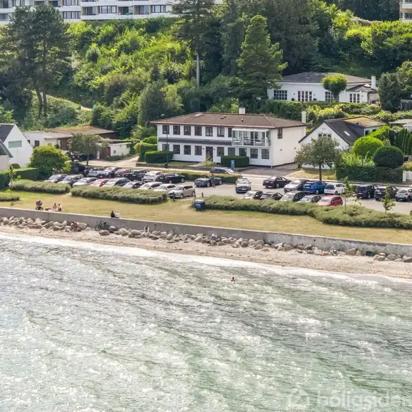 Bygning med parkeringsplads omgivet af biler ved kystlinje; græsplæne og strand foran, omgivet af flere huse og frodig skov i baggrunden.