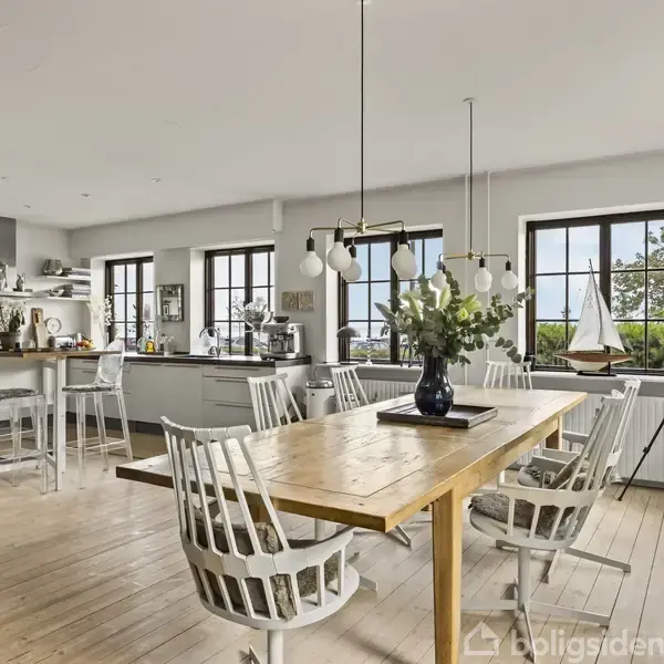 Et stort, lyst køkken med hvidt interiør, hvor et træbord med hvide stole står central. På bordet er en vase med blomster. Området har store vinduer og et teleskop.