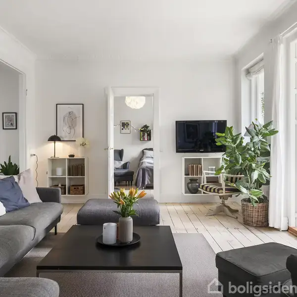 Gråt sofaarrangement og sofabord med blomster. TV monteret på væggen. Store vinduer og dør til terrasse, lyseblå farvet vægge, trægulve, grønne planter og kunstværker på væggene.
