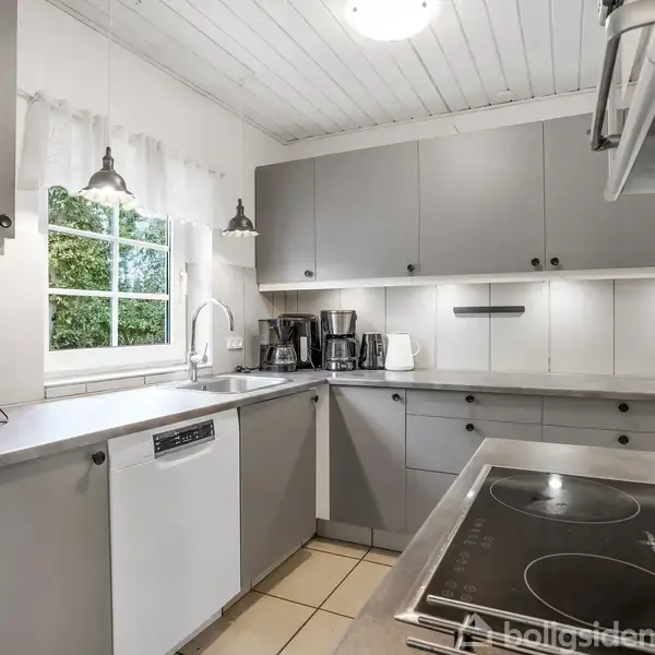 Køkken med grå skabe, står stille under hvidt loft. Køkkenbordet har kaffemaskine, brødrister og vask ved et vindue. Kogeplade og ovn er synlige på højre side.
