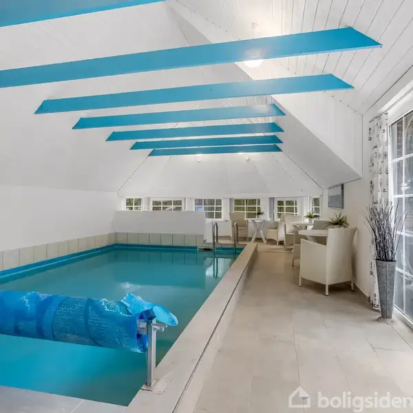 En indendørs swimmingpool er stille i et lyst rum med blå bjælker. Store vinduer til højre viser en grøn have udenfor. I baggrunden står hvide stole og planter.