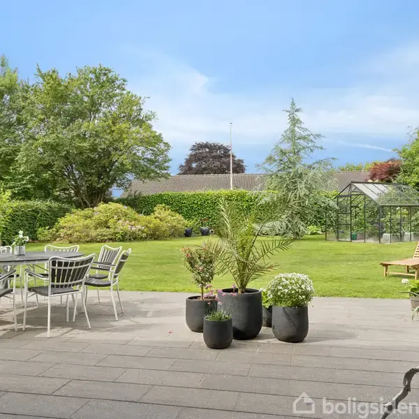 En terrasse har bord og stole, omkranset af grønne planter i krukker. I baggrunden ses en velplejet have med buske, træer og et drivhus.