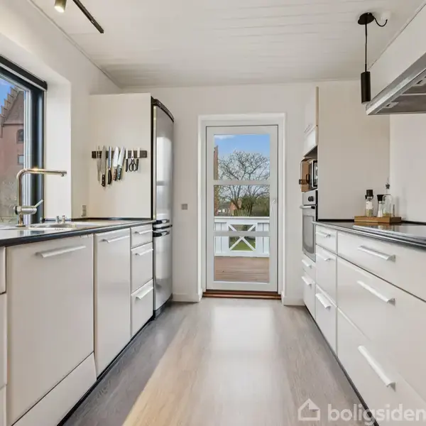 Et moderne køkken med hvide skabe og sorte bordplader strækker sig langs væggene. Et vindue og en glasdør giver udsigt til en balkon med træer udenfor.