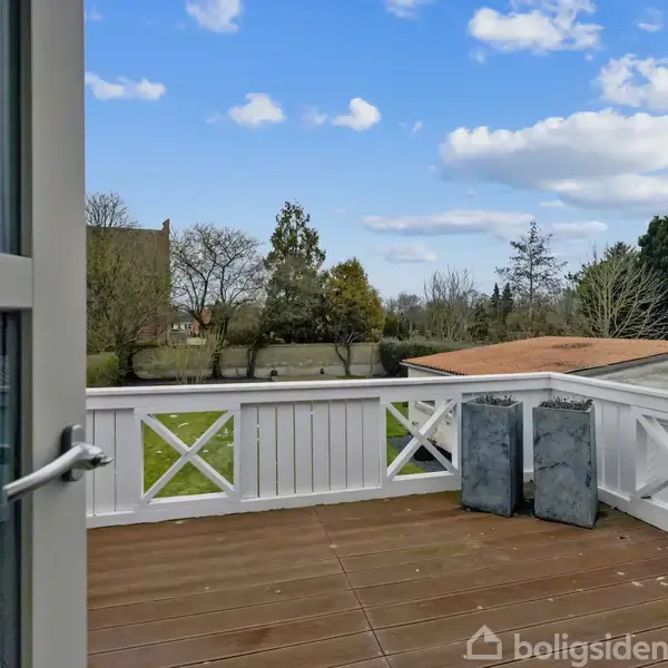 Dør åbner ud til en træterrasse med to kvadratiske plantekasser. I baggrunden ses en have med græs og træer under en blå himmel med skyer.