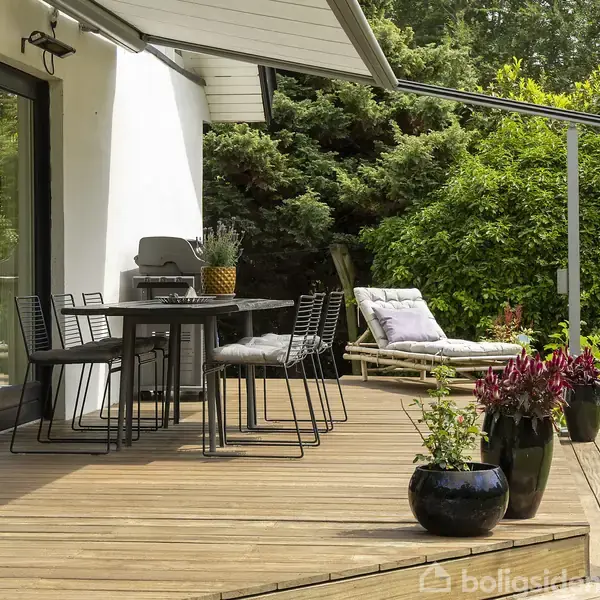 Terrassemøbler står på en træterrasse; sorte stole om et bord. Bagved er en liggestol med pude. Terrassen grænser op til grøn beplantning, og der er blomster i krukker.