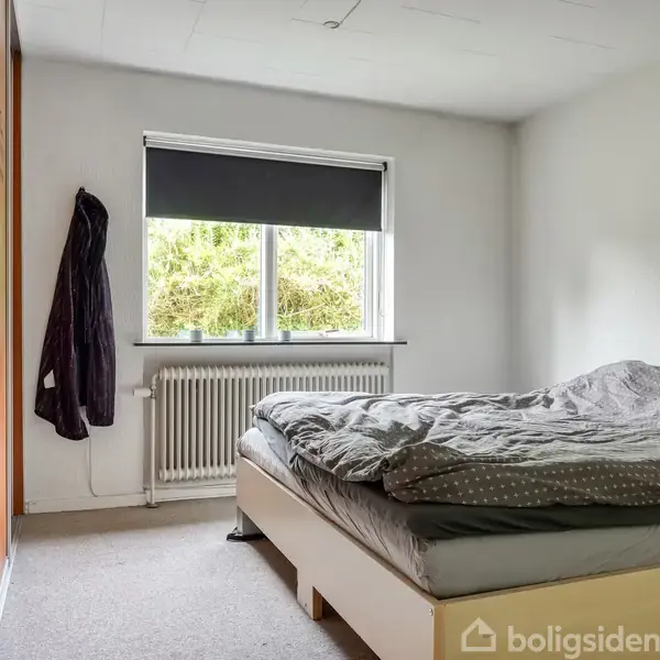 Soveværelse med en dobbeltseng dækket af gråt sengetøj. En væg har et vindue med udsigt til træer og en radiator under. En kappe hænger på skabsdøren.