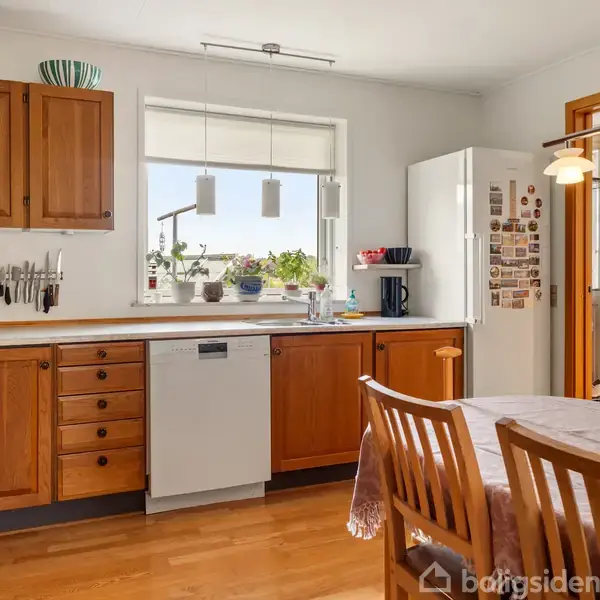 Et køkken med træskabe og hvid opvaskemaskine ved siden af vasken. Køleskabet har magneter og billeder. En træbord med stole står i forgrunden. Vindue med udsigt i baggrunden.