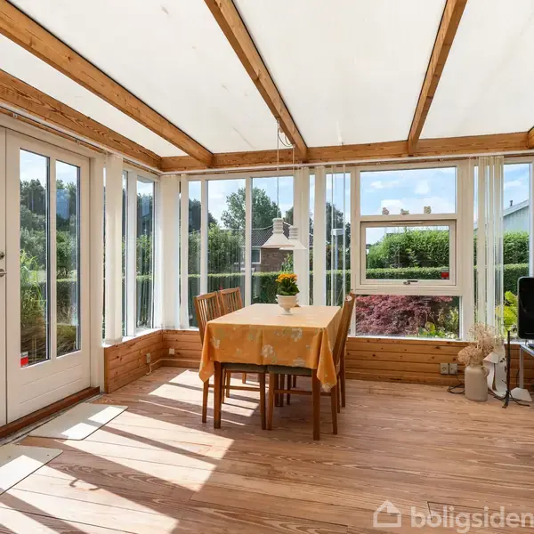 Et bord med to stole står i en lys udestue. Sollys strømmer gennem store vinduer, omgivet af grønne planter udenfor. En lille pejs kan ses gennem vinduet.