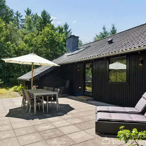 Sort hus med terrasse har spisebord under parasol og liggestol. Omgivet af grønt løv og træer i baggrunden. Sollys skaber en hyggelig atmosfære.