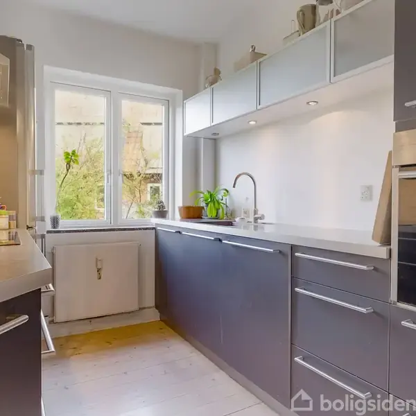 Et moderne køkken med grå skabe og skuffer. En ovn er indbygget i skabsvæggen. Et vindue over køkkenbordet kaster lys ind. Planter og krydderier står på bordet.