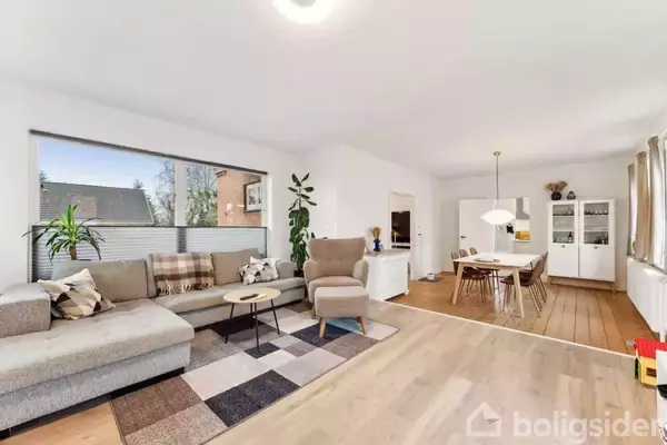 Grå sofa med tæppe og puder står i en lys stue. Ved siden af er en lænestol, spisebord og reol. Store vinduer lader dagslys ind, og der er planter.