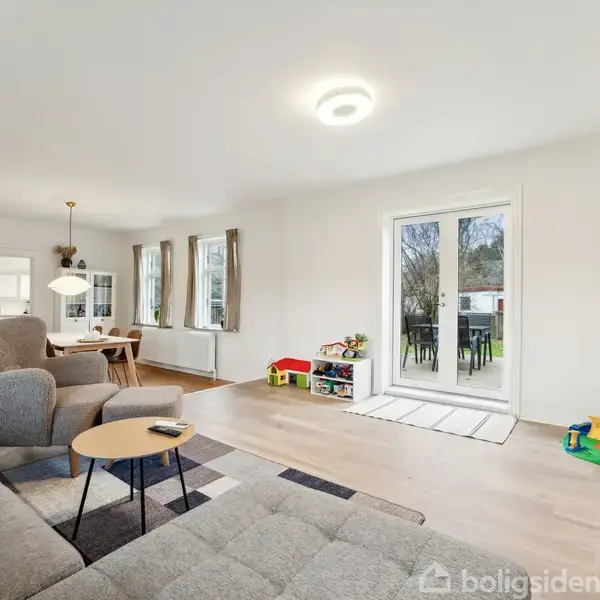 En stue med en grå sofa og lænestol står i rummet. Et sofabord er foran sofaen. I baggrunden ses spisebord ved vinduer. Legetøj er på gulvet nær terrassedøren.