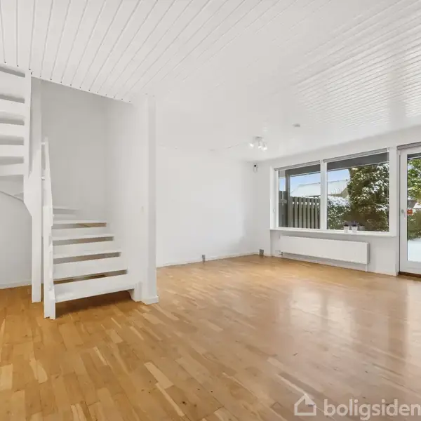 Trappe med hvide trin og gelænder fører opad. Stuen har trægulv, store vinduer og en dør fører ud til en have med træer. Ingen tekst.