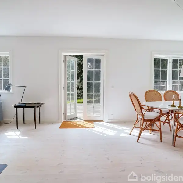Stue med lyse vægge har et blåt sofaarrangement til venstre og et spisebord med flettede stole til højre. Franske døre fører ud til en have.