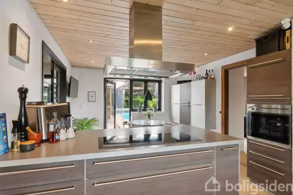 Køkken, med kogeplade og emhætte, vender mod en lys stue med store vinduer, der kigger ud på en have med pool. Peberkværn og madlavningsartikler står på bordet.