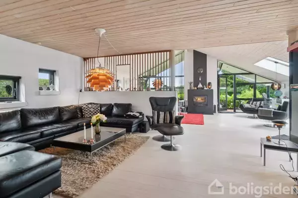 Lædersofa med sofa- og lænestol-arrangement i moderne stue, der rummer pejs. Stort vinduesparti giver udsigt til frodig have. Loftet er beklædt med lyse træplanker. Skillevæg med trælameller.