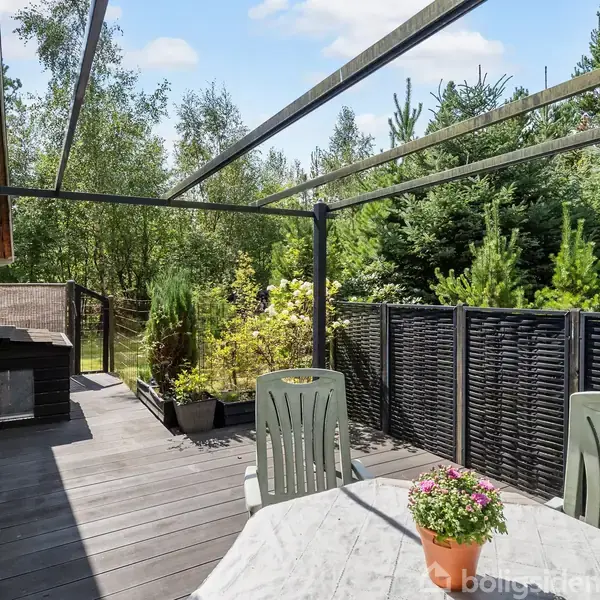 Terrassebord med blomsterkrukke står på trædæk. Plaststole er omkring bordet. Hegn og buske omkranser området, mens skoven er synlig i baggrunden under en delvist overskyet himmel.