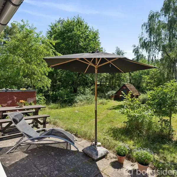 Liggestol og parasol står på en terrasse med bordbænk. Terrassen er omgivet af græsplæne og træer på en solrig dag. En lille træhytte ses i baggrunden.