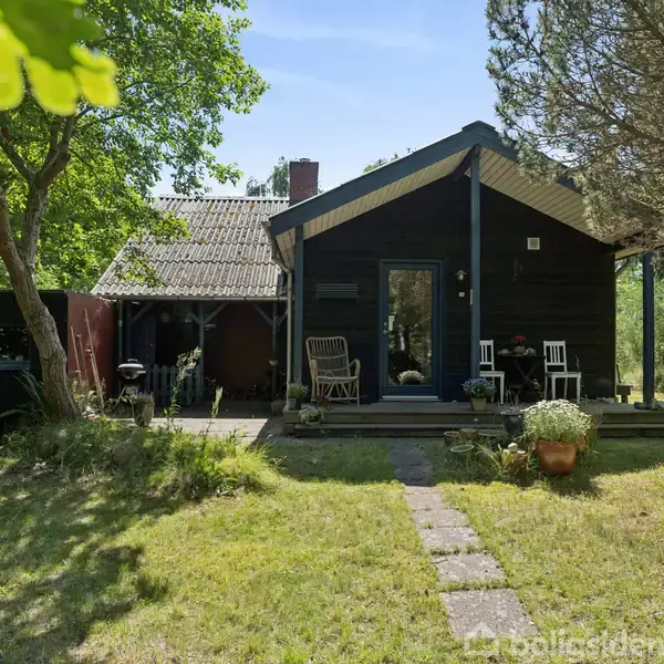 Et træhus står i en have med græsplæne og omgivne træer. Foran huset er der en terrasse med stole og bord, hvor der er placeret blomsterkrukker.
