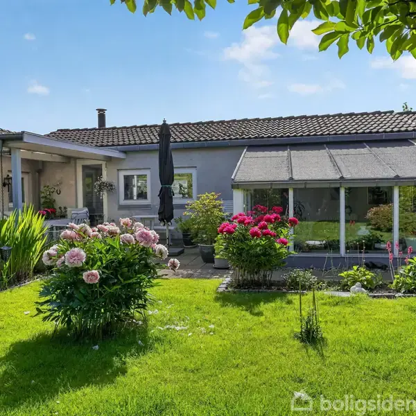 Have med blomstrende buske og grøn græsplæne foran et hus med fladt tag og udestue. En stor parasol står ved et bord i haven under en klar blå himmel.
