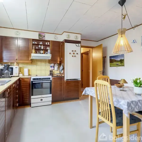 Køkken med brune skabe og hvidevarer, herunder ovn og køleskab. Et spisebord med fire stole står til højre, under en moderne lampe. Borders dekoration med planter og maleri.