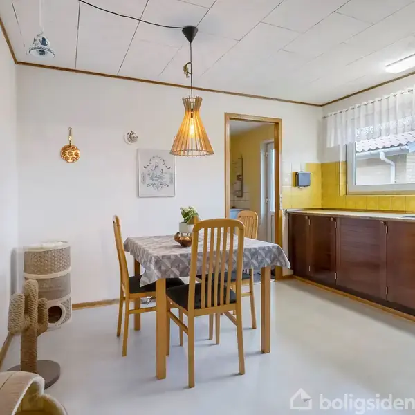 Spisebord med dug står centralt i et køkken med gule fliser og brunt skab. To stole om bordet. Kattelegetøj står ved væggen. Hängebelysning lyser rummet op.