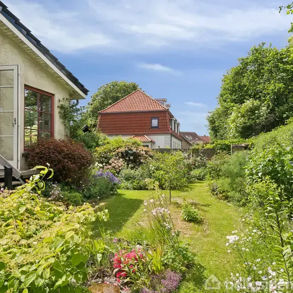 Hus med åbne havedøre, omgivet af en frodig have med blomster og grønne buske. I baggrunden ses et rødt tegltag og blå himmel. Ingen tekst i billedet.