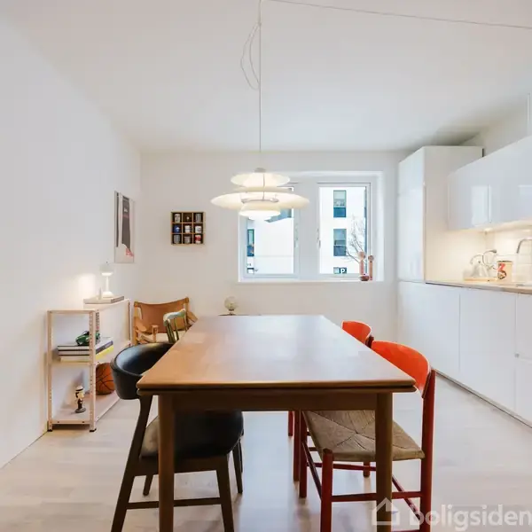 Et spisebord med stole står centralt i et lyst, moderne køkken. Over bordet hænger en lampe. Køkkenet har hvide skabe, en ovn, og et vindue med udsigt til bygninger.