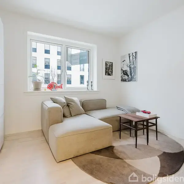 Et spisebord omgivet af stole står i et lyst køkken. Der ses moderne hvide skabe, et komfur og en ovn. Til venstre er et vindue, til højre en døråbning.