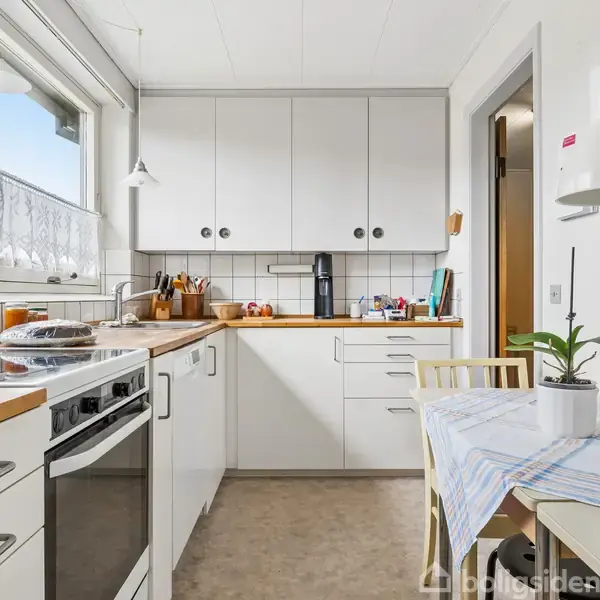 Et lyst køkken med hvide skabe og træbordplade. En komfur med en kande står til venstre, mens et bord med en plante under en lampe står til højre.