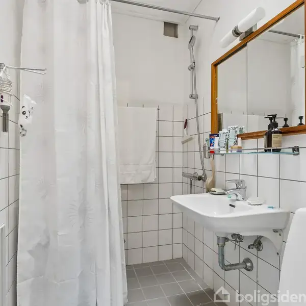 Et badeværelse med hvid-flisebelagte vægge. Der er en vask med spejl over, en bruser med forhæng til venstre, og en radiator. Hylde over vasken med toiletartikler.