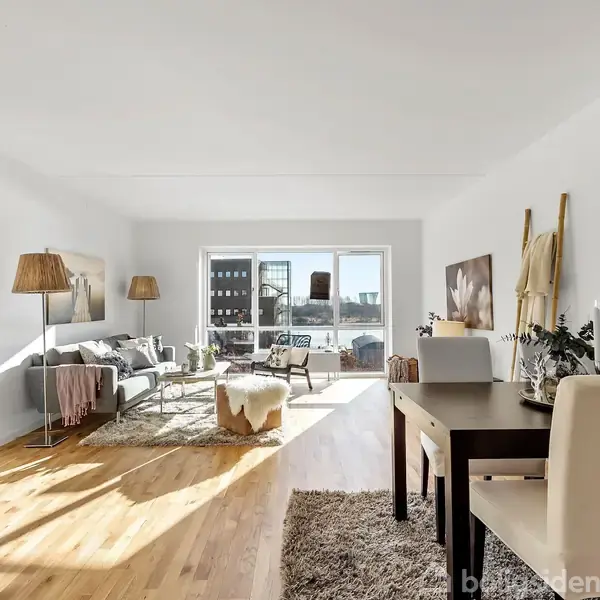 Stue med sofa og lænestole arrangeret omkring et sofabord, belyst af naturligt lys fra store vinduer. Et spisebord med stole står til højre, omgivet af dekorative elementer.