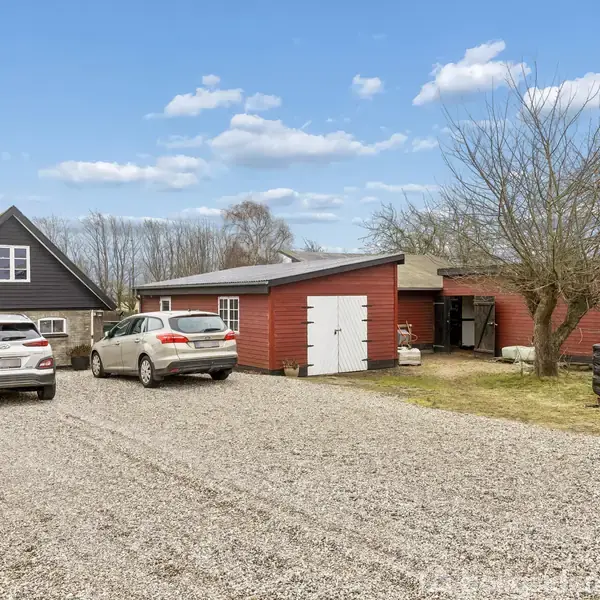 To biler står parkeret foran et hus med rødt udhus. Grusvej omgiver bygningerne, og nogle træer ses i baggrunden under en blå himmel med lette skyer.