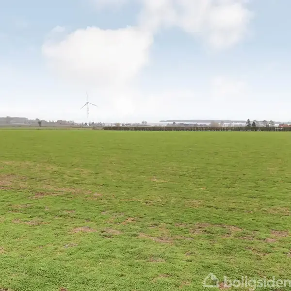 Grøn mark strækker sig mod horisonten, hvor en vindmølle står stationært, omgivet af marker og få bygninger under en lys, blå himmel.