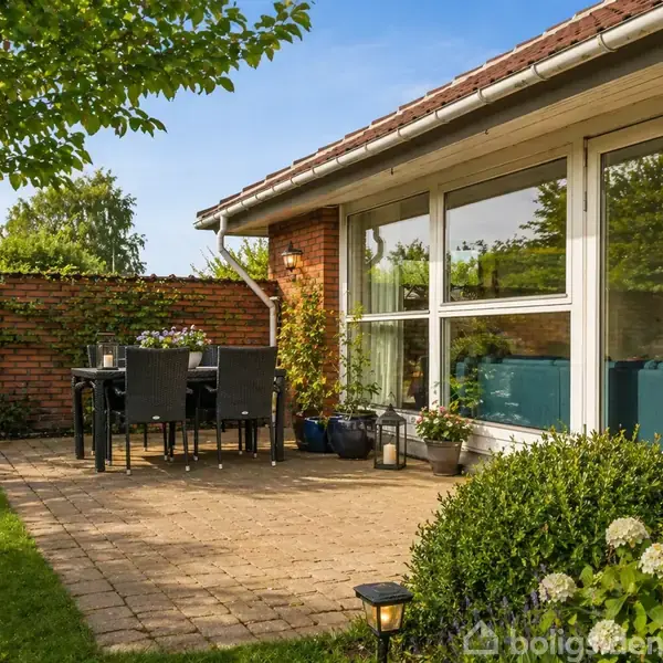 Terrasse med bord og stole placeret på flisebelagt område foran et hus. Huset har store vinduer, og omgivelserne inkluderer grønne planter og en murstensvæg.