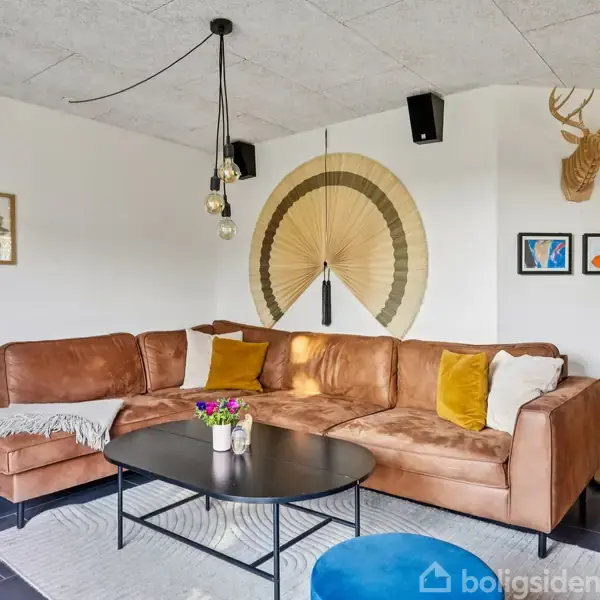 Brun hjørnesofa med gule og hvide puder står i en moderne stue. Sofaen står ved et hvidt vægpanel med dekorationer. Foran står et ovalt, sort sofabord på et lyst tæppe.