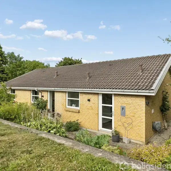 Huset, lavet af gule mursten, står stille omkranset af en velholdt have med grønne planter og en græsplæne. Himlen er klar med få skyer, der skaber en fredelig atmosfære.