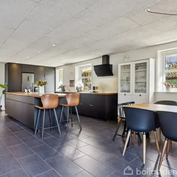 Et moderne køkken med mørke skabe og en stor ø. Der står tre høje stole ved øen, og et spisebord med seks stole er placeret ved siden af vinduerne.