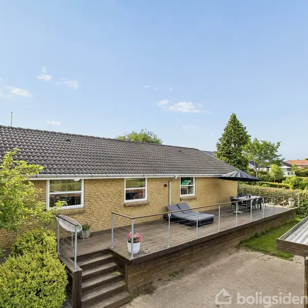 Et gult murstenshus med mørkt tag har en stor terrasse med liggestole og planter. Omgivet af grønne træer og buske med en carport til højre i billedet.