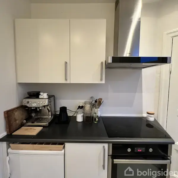 Køkkenbord med espressomaskine og skærebræt står ved siden af en kogeplade med køkkenredskaber og skab ovenover. Til højre er der en emhætte og en lukket dør.