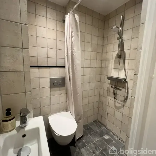Brusekabine med moderne bruser i klinisk flisebadeværelse. Toilet ved siden af, adskilt af hvidt bruseforhæng. Dispenser og vandhane ses på kanten af vasken.